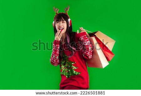Happy pretty asian woman in red christmas theme cloth wearing reindeer headband and earmuffs carrying paper shopping bags posing on green screen background. Merry Christmas concept.