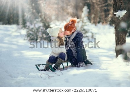 A beautiful little girl is sitting on a sled in a winter park. A girl in a white fur coat in a winter snowy forest on a sled. It's snowing, children on sleds are kissing.