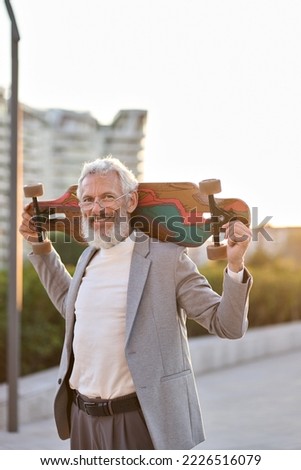 Active smiling happy cool gray haired bearded old senior business man skater wearing suit holding skateboard standing in city on sunset outdoors. Older people freedom spirit concept. Vertical portrait