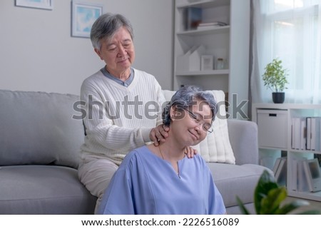 Asian old senior elderly wife sitting on floor having problem with suffer neck painful shoulder while care husband standing behind help massaging back.