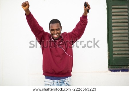 Portrait of excited young african man enjoying listening to music with earphones and dancing against wall outside