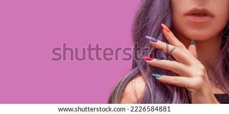 Female hand with colorful nail design. Glitter nail polish manicure: green, pink, purple and orange. Female model hand with perfect colorful manicure touch her long hair on pink background. Copy space