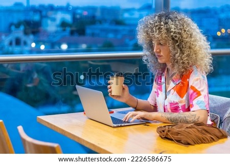 Portrait of a smiling young Caucasian woman with curly blonde hair, working in a coffee shop with her laptop and smart phone