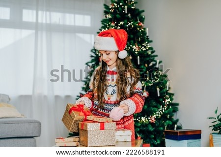 A happy smiling little girl in Santa hat and mom's festive knitted sweater packing gift boxes on the Christmas tree background. Christmas mood concept. Preparing or opening presents. Selective focus.