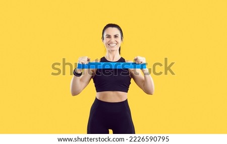 Cheerful sports woman on yellow background with her hands stretches rubber elastic band for training. Portrait of beautiful smiling woman with slender body who is wearing black sportswear set.