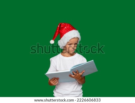 Boy in Santa Claus hat is thinking and looking at an open book. Green background with space for text. Selective focus. Picture for articles and advertisements about children, holidays, Christmas.