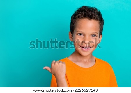 Photo of pretty sweet little guy dressed orange t-shirt pointing thumb empty space isolated teal color background