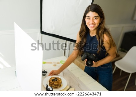 Cheerful latin photographer making eye contact while holding her professional camera and doing food styling during a shooting