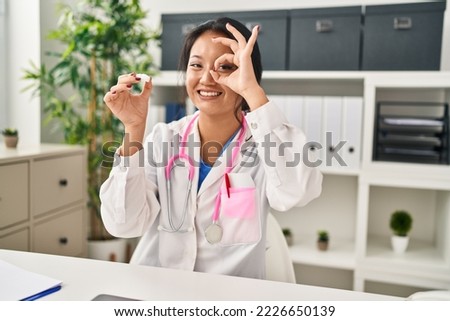 Young asian woman wearing doctor uniform and stethoscope smiling happy doing ok sign with hand on eye looking through fingers 