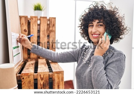 Young middle east woman business worker talking on smartphone writing on cork board at office