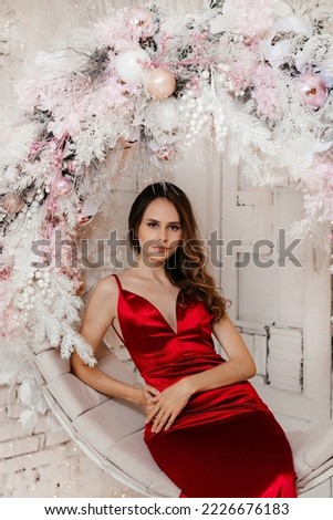 a girl in a tight-fitting red silk dress sits in a New Year's round hanging composition decorated with white and pink Christmas balls and white artificial leaves