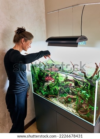 A beautiful smiling woman stands with scissors and looking into aquarium aquascape. Trimming concept. Royalty-Free Stock Photo #2226678431