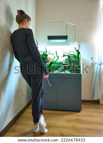 A woman standing near the wall in the room and holding scissors in the hand. Beautiful planted aquarium at the background. Aquascape trimming concept.