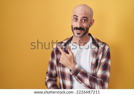 Hispanic man with beard standing over yellow background cheerful with a smile of face pointing with hand and finger up to the side with happy and natural expression on face 