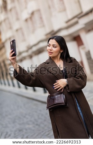 young brunette woman in brown coat taking selfie on blurred street in prague