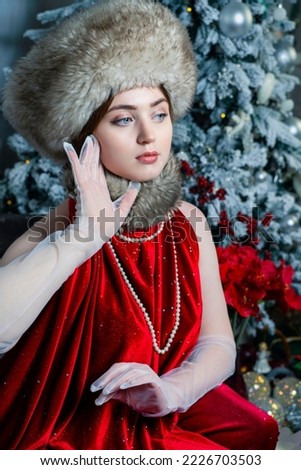 A beautiful woman in a fur hat, fur collar and pearls on the background of a Christmas tree. Russian style.