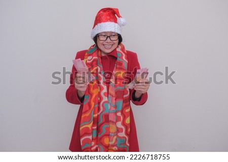 A middle-aged Asian woman wearing a red coat, a Santa Klaus hat and shawl holds some money in both hands; excited, happy expression.