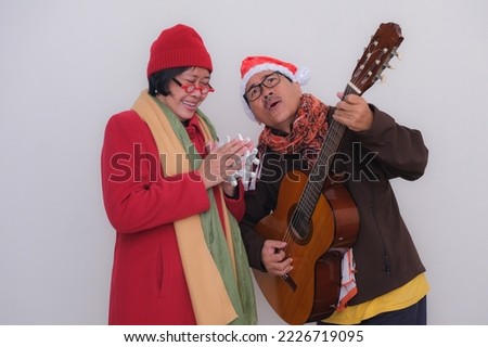 A husband wearing a dark coat and Santa Klaus hat plays guitar for his smiling wife holding a christmas ornament.