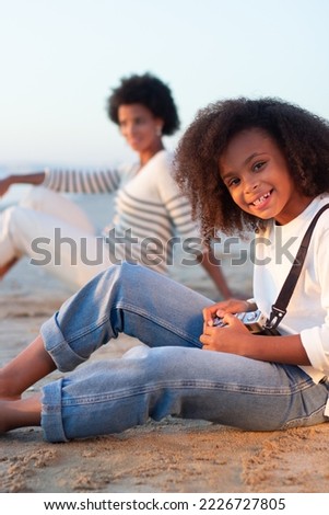 Smiling girl with old-fashioned camera. Female model with curly hair and in white pullover holding camera on beach. Mother in background. Childhood, hobby concept