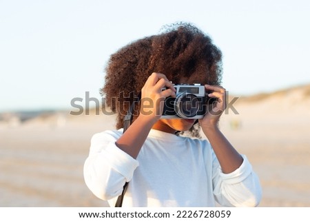 Portrait of little girl with old-fashioned camera. Female model with curly hair and in white pullover holding camera on beach. Childhood, hobby concept