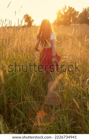 With her back to camera, long-haired girl dressed as hippie walks through field at sunset. copy space