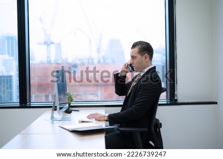 Business man using smartphone contact customer at desk on office in city with high building view, serious and intense emotion business onwer sitting beside glass window.