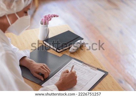 Side view of female doctor at work during pandemic. Woman in medical uniform and protective mask sitting at table filling application form with pen. Doctors job, health care, medical service concept