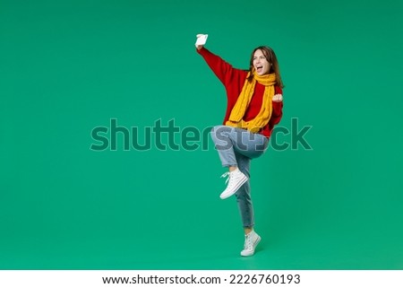 Full length side view of happy young brunette woman 20s in knitted red sweater yellow scarf doing selfie shot on mobile phone doing winner gesture isolated on green color background studio portrait