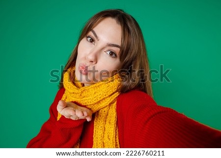 Close up of pretty young brunette woman 20s in knitted red sweater yellow scarf standing doing selfie shot on mobile phone blowing sending air kiss isolated on green color background studio portrait