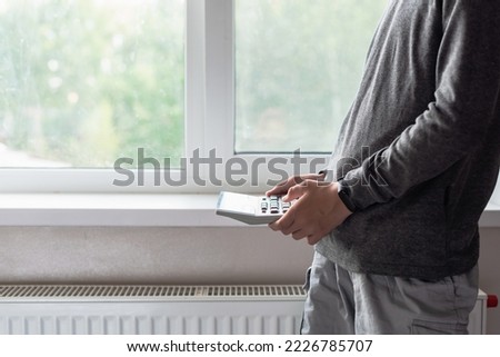 Hands with calculator near a heating radiator. Natural gas consumption, cost of utilities and saving concept. Heating season, comfortable living conditions, energy crisis, gas crisis, saving energy Royalty-Free Stock Photo #2226785707