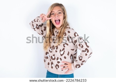 caucasian teen girl wearing animal print sweater over white background making v-sign near eyes. Leisure, coquettish, celebration, and flirt concept.