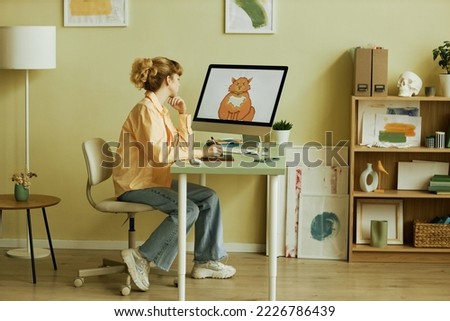 Young creative blond female graphic artist in casualwear sitting by workplace in front of computer screen with drawn picture of cat