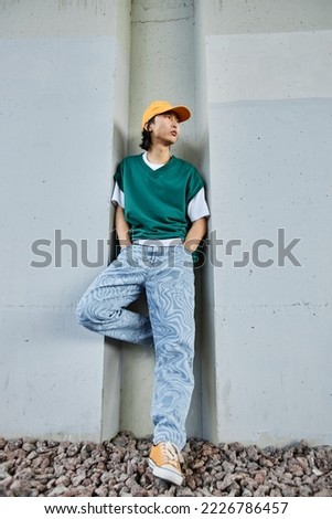 Vertical portrait of young Asian man wearing colorful street style clothes standing by concrete wall in urban setting Royalty-Free Stock Photo #2226786457