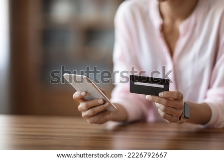 Unrecognizable woman in pink blouse sitting at desk, holding modern cell phone and plastic credit card, lady shopping online or paying bills on Internet, cropped, copy space. Online banking concept Royalty-Free Stock Photo #2226792667