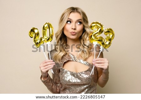 Charming young blonde girl with curly hair wearing sparkle dress holding balloons number 2023 in her hands on a beige background, new year holiday concept Royalty-Free Stock Photo #2226805147