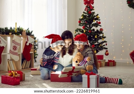Happy family discovers wonderful surprise while opening Christmas presents. Mother and two children in red Santa hats looking inside gift box sitting together on floor in beautiful festive living room