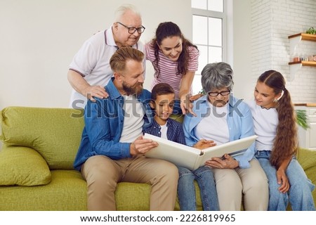 Lovely happy smiling big family with parents, kids, grandparents looking at family photo album together sitting in modern light living room at home. Emotional family moments, togetherness concept.