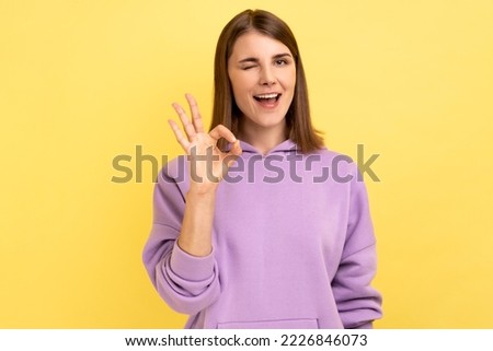 Woman with toothy smile, winking to camera and showing thumb up, approval sign, satisfied with service, good feedback, wearing purple hoodie. Indoor studio shot isolated on yellow background.