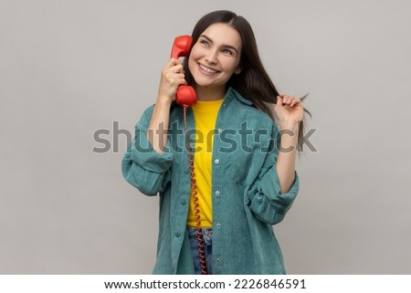 Smiling woman talking via red retro landline phone, having pleasant conversation, looking away with smile, touching hair, wearing casual style jacket. Indoor studio shot isolated on gray background. Royalty-Free Stock Photo #2226846591