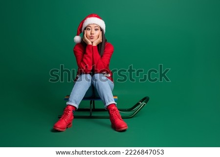 Full length photo of flirty girl dressed red x-mas pullover sending kiss riding sledge arms cheeks empty space isolated green color background