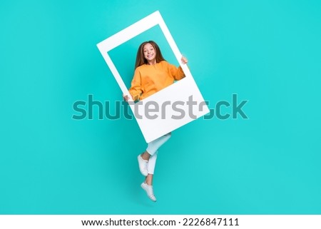 Full length photo of charming dreamy schoolgirl wear orange sweatshirt jumping high holding white retro frame isolated turquoise color background