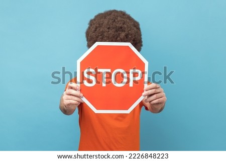 Portrait of man with Afro hairstyle wearing orange T-shirt covering face with Stop symbol, anonymous person holding red traffic sign, warning to go. Indoor studio shot isolated on blue background.