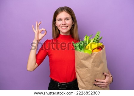 Young English woman holding a grocery shopping bag isolated on purple background showing ok sign with fingers