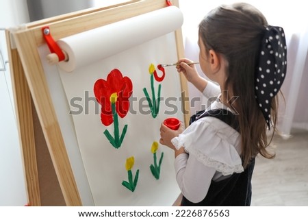 A little girl with a palette in her hands draws flowers on an easel.