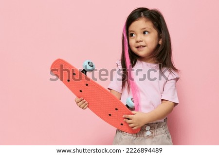 a cute little girl is standing on a pink background in a pink T-shirt, holding her skate in her hands, looking at it from all sides and smiling sweetly while hugging it to her