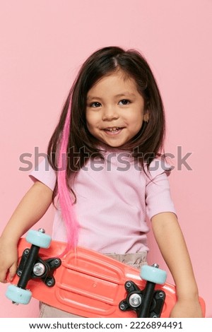a cute little girl is standing on a pink background in a pink T-shirt, holding her skate in one hand in front of her, and raising the other behind her head