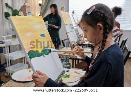 A little girl concentrates on acrylic color picture painting on canvas with student children in an art classroom, creative learning with talents and skills in the elementary school studio education. 