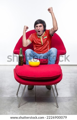 Soccer fan man with red jersey and face painted with the flag of the IR IRAN team screaming with emotion on Red armchair from your house with snack and beer. Royalty-Free Stock Photo #2226900079