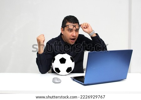 Latino adult office man watches football games on his work laptop during office hours in the morning, he sees him excited, nervous, surprised next to his soccer ball
