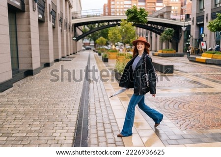 Full length of positive redhead woman wearing fashion hat walking on European city street holding in hand closed transparent umbrella. Concept of modern female lifestyle at autumn season. Royalty-Free Stock Photo #2226936625